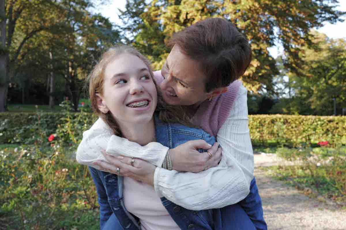 Mutter umarmt ihre Tochter von hinten, beide lachen, Herbst im Park