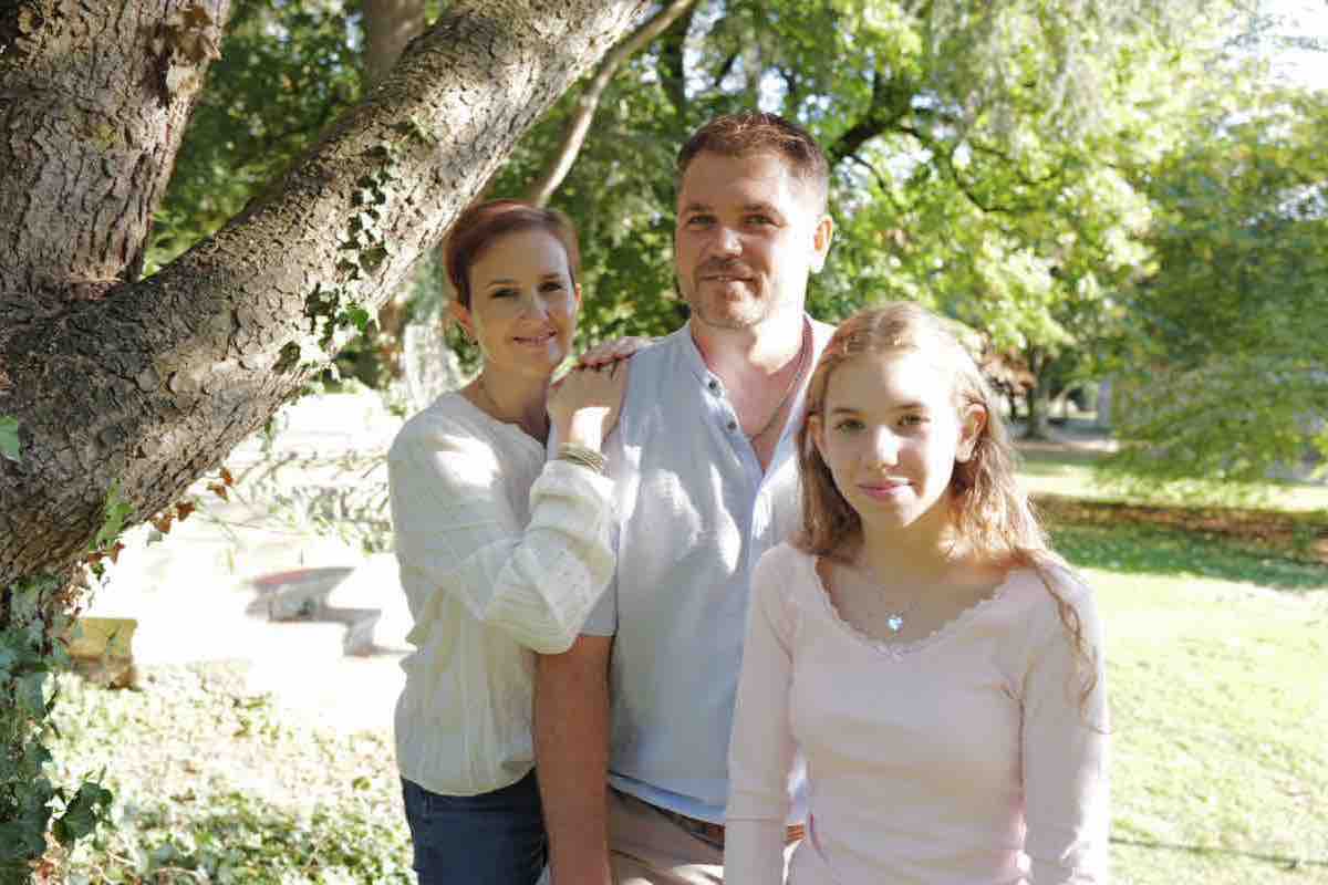Familie an einem Baumstamm, die Mutter legt die Hand auf die Schulter des Vaters